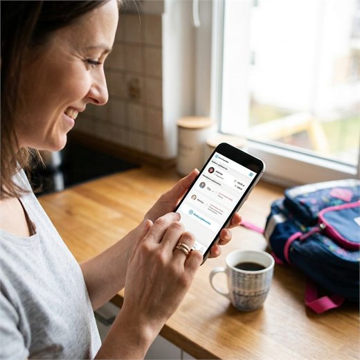 Uśmiechnięta mama w kuchni zamawia obiady dla dziecka w aplikacji e-Stołówka na telefonie